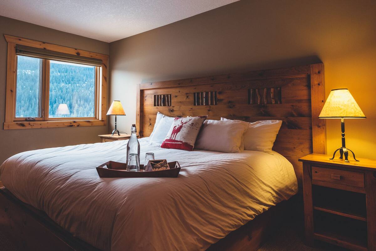 A bedroom with a large bed, wooden headboard, two bedside lamps, a nightstand, and a window with a view of a forest.