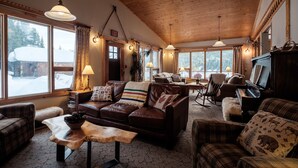 Una sala de estar acogedora con paneles de madera, sofás de cuero, un piano y ventanas amplias que ofrecen vistas nevadas.