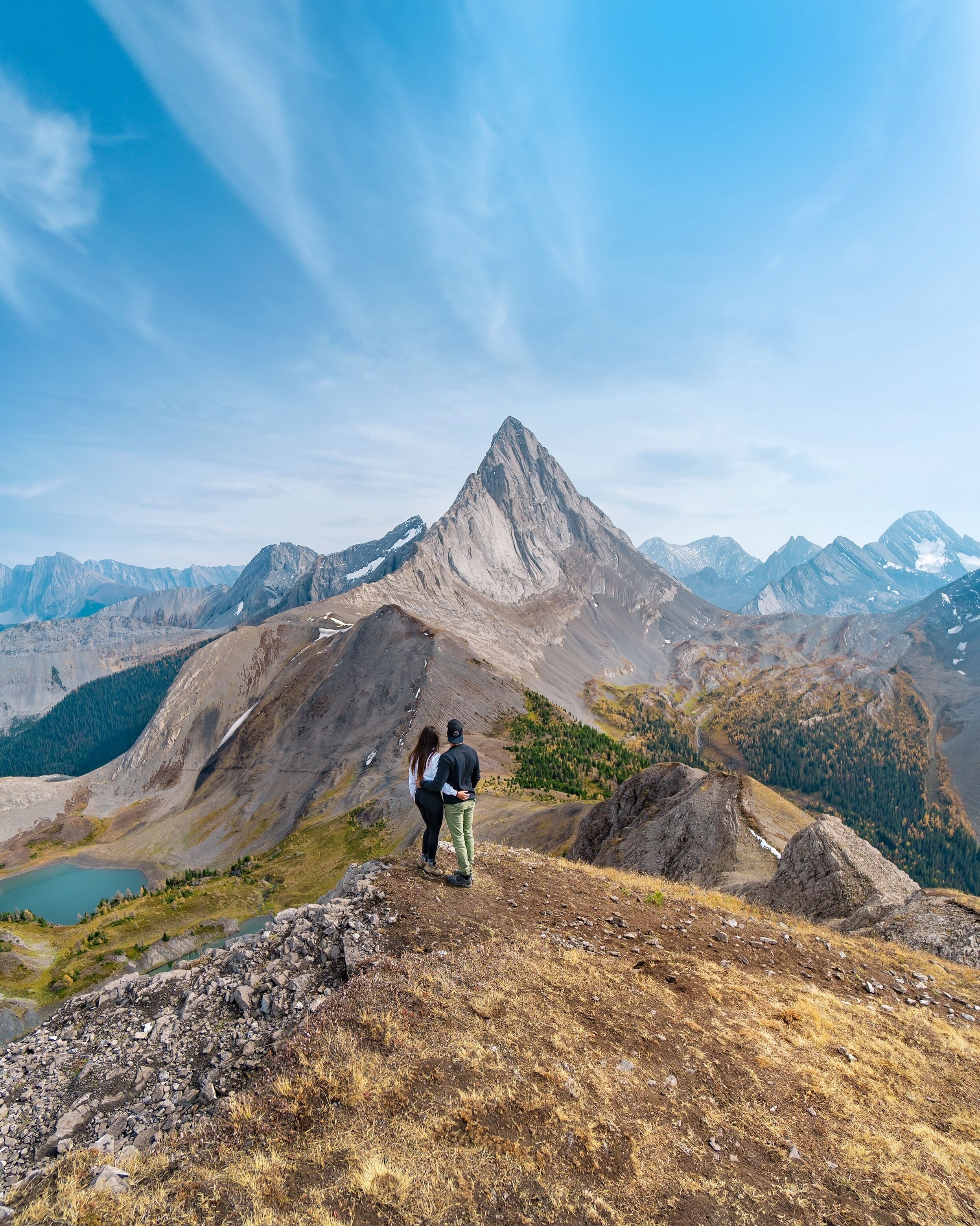 A couple standing on a mountain ridge overlooking a valley with a lake and distant mountains.