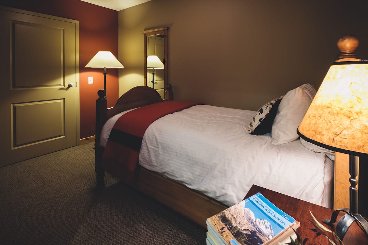 A hotel room with a bed, two bedside lamps, a nightstand with a book, and a door.