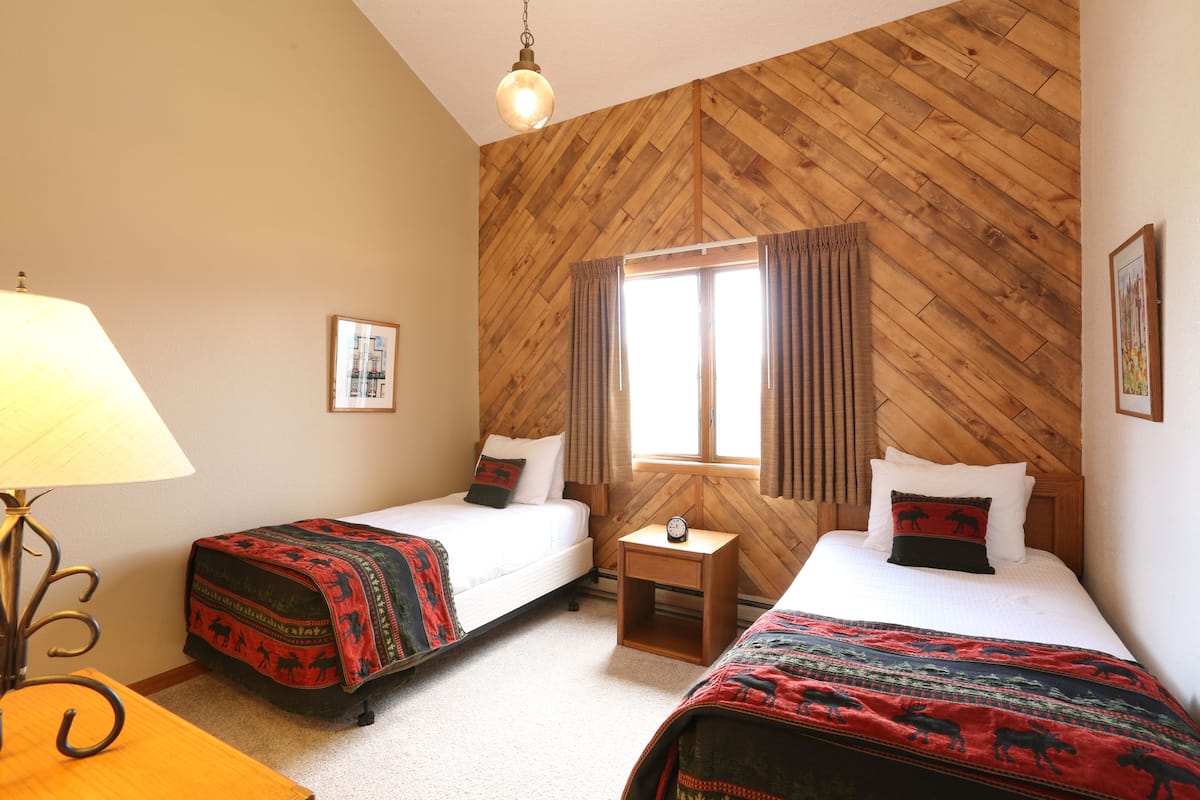 A room with two beds, wooden walls, a window with curtains, a bedside table with a clock, and a lamp.