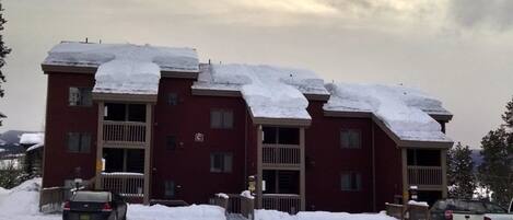 A multi-story building with snow-covered roofs and a parking area with several vehicles.
