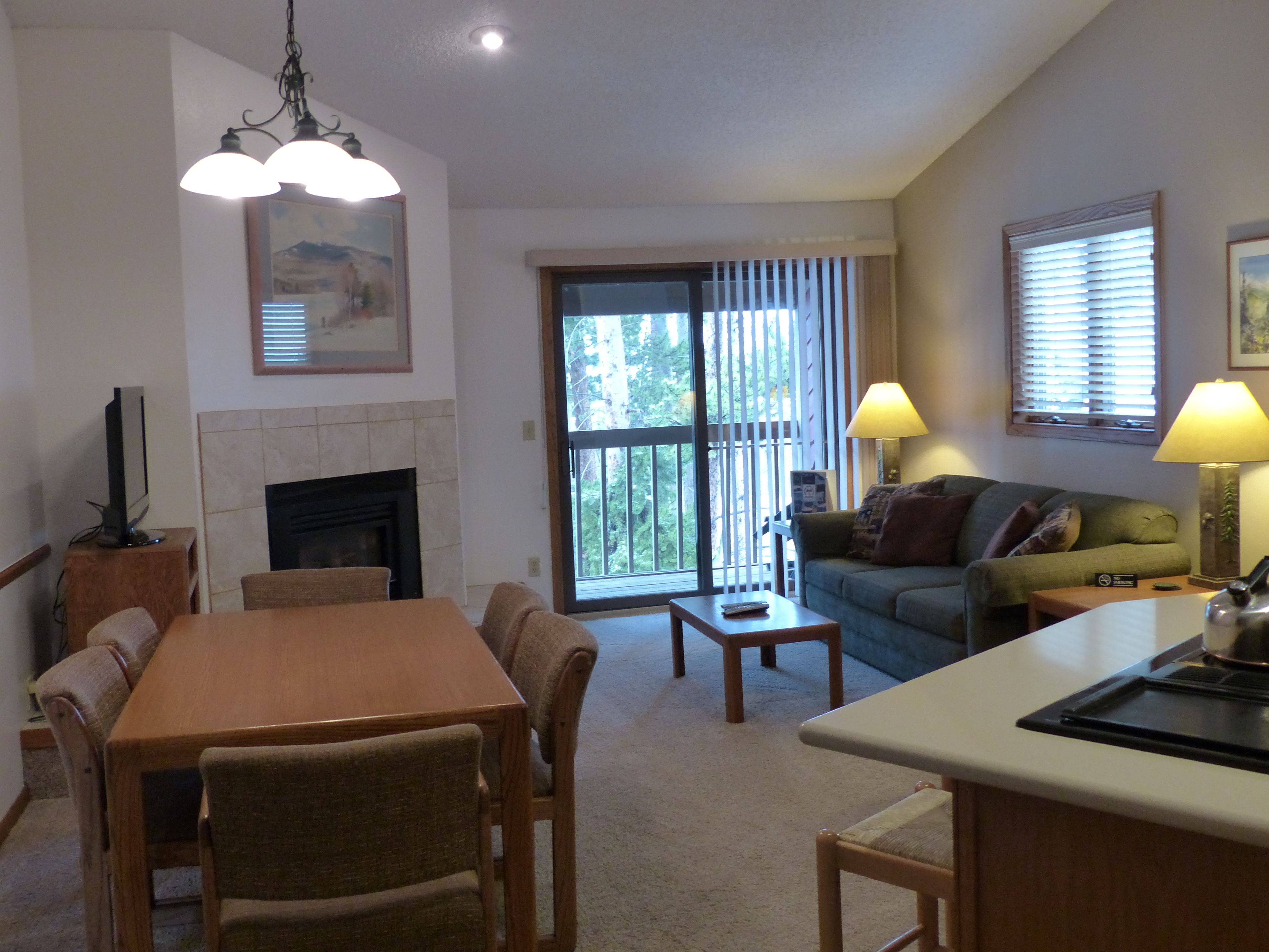 A living room with a fireplace, a dining table with chairs, a sofa, and a kitchen area with a counter and a lamp.