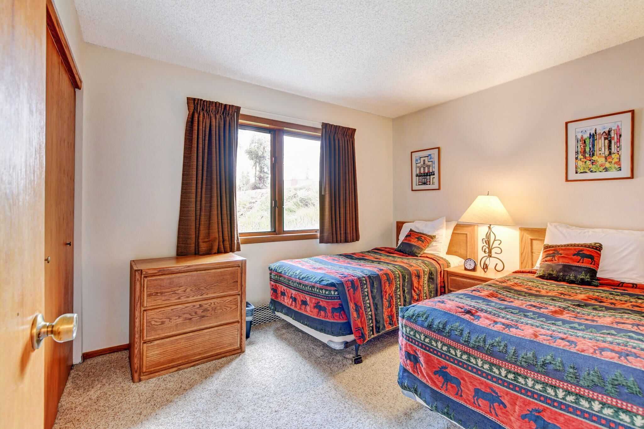 A room with two beds, a wooden dresser, a window with curtains, a nightstand with a lamp, and two framed pictures on the wall.