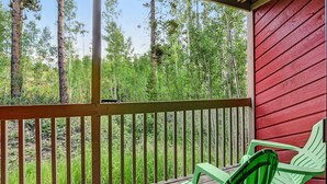 A wooden deck with a red wall and green chairs, overlooking a forested area.