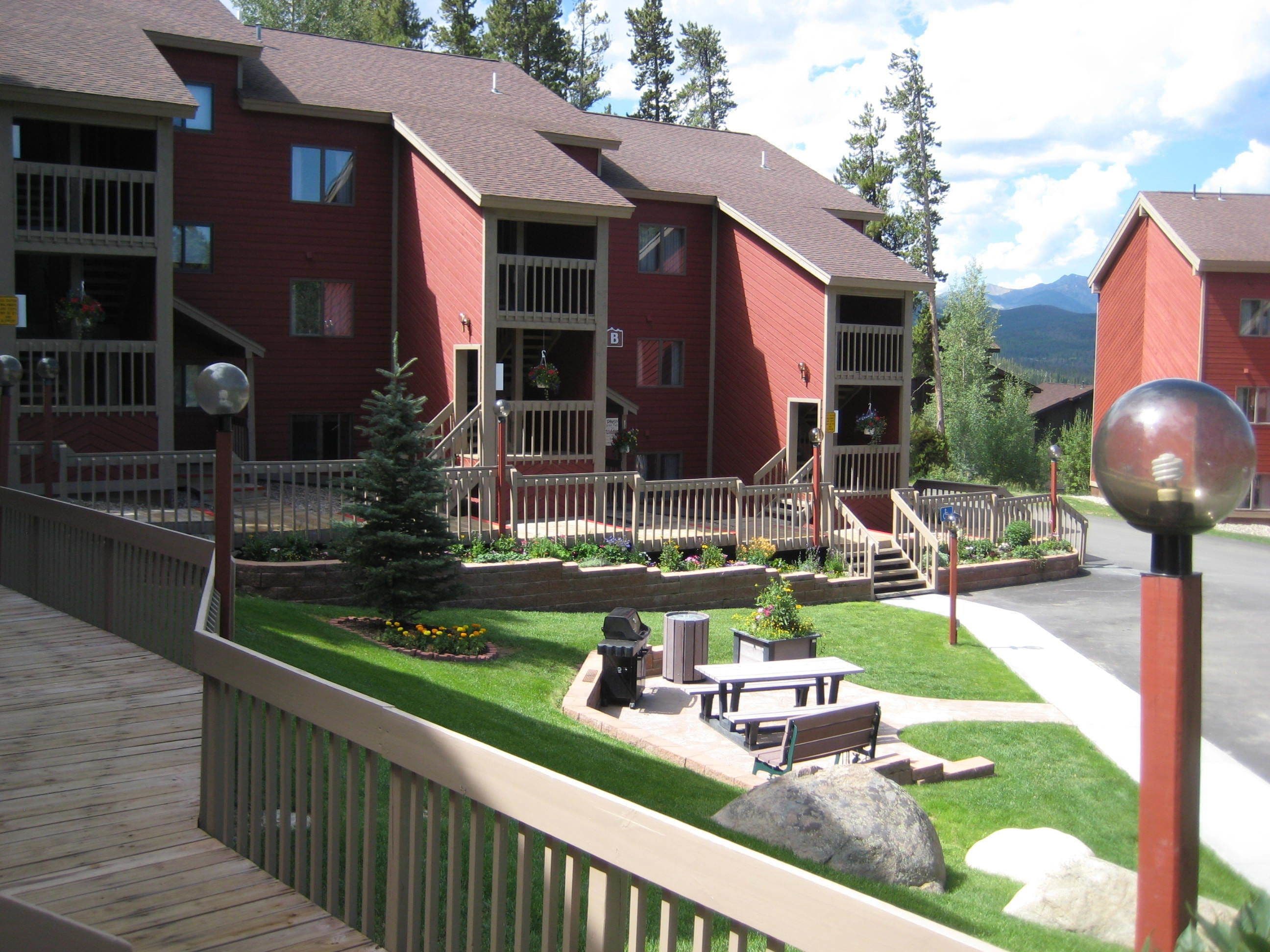 A red multi-story building with balconies, a small lawn, picnic tables, and a wooden deck.