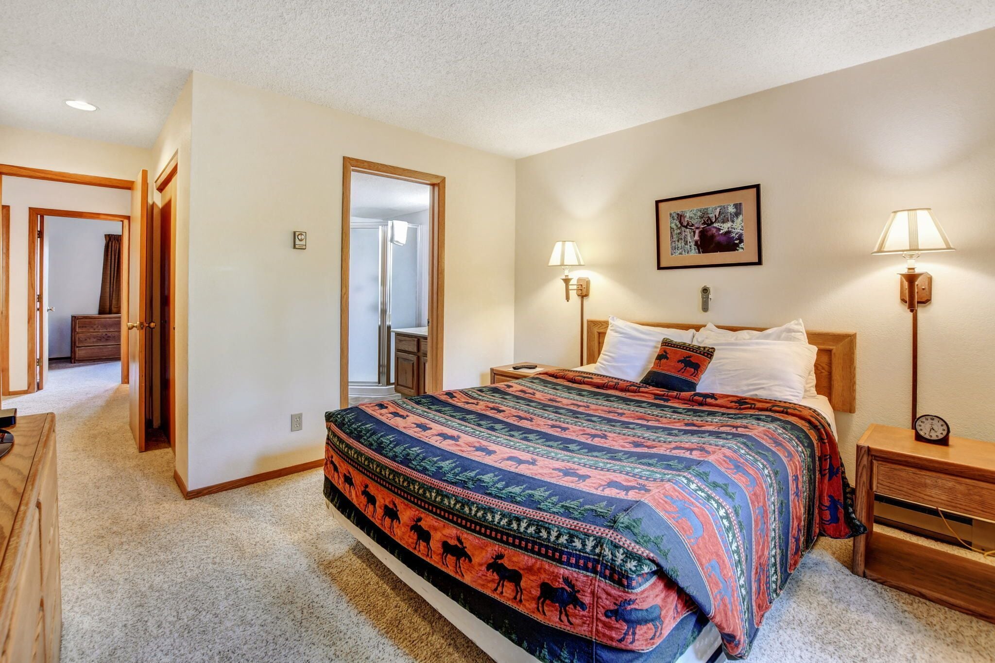 A bedroom with a large bed, two bedside lamps, a nightstand, a wooden dresser, and a door leading to another room.