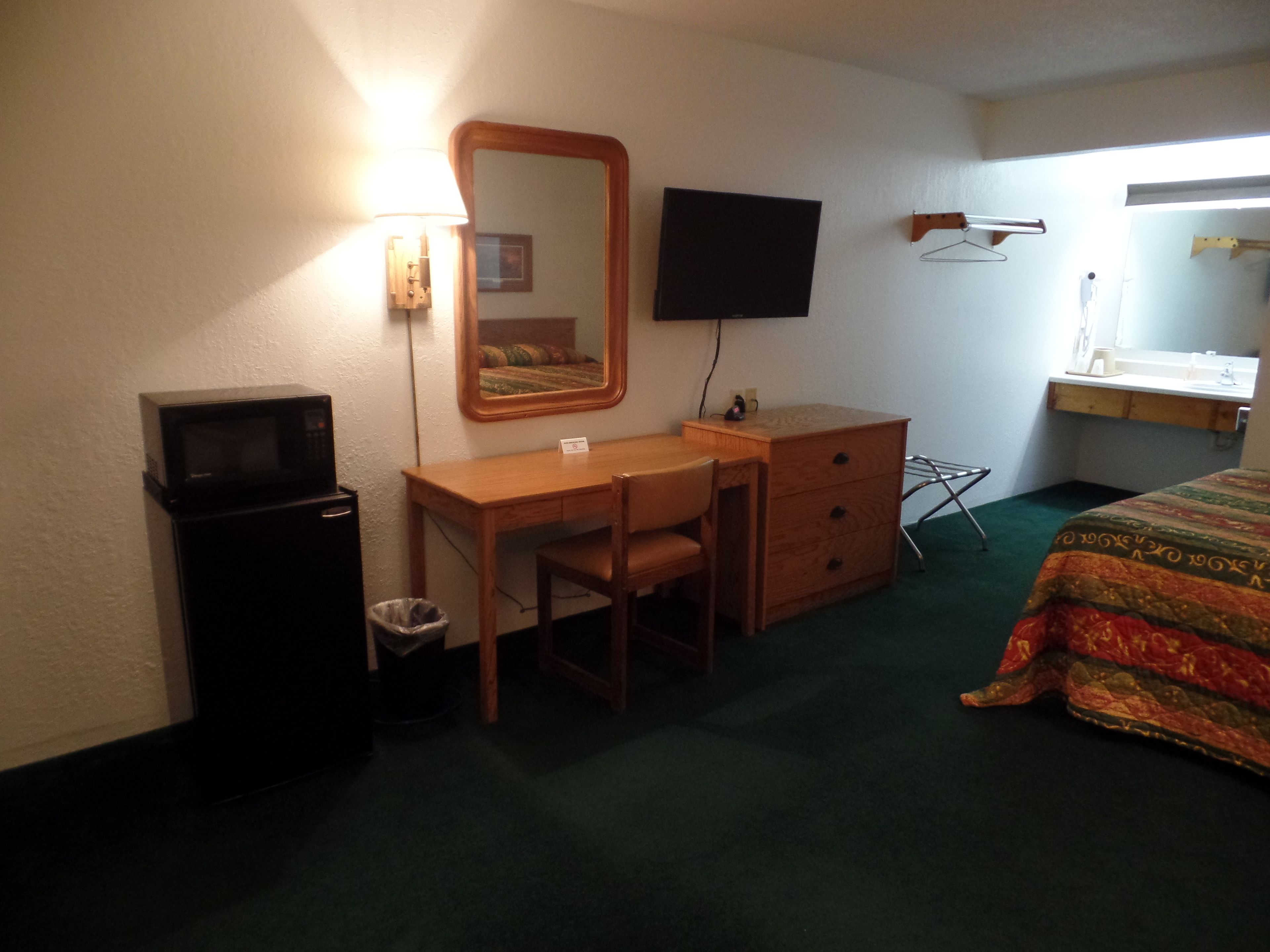 A hotel room with a desk, chair, and a bed. There is a television mounted on the wall, a mirror above the desk, and a small trash bin.