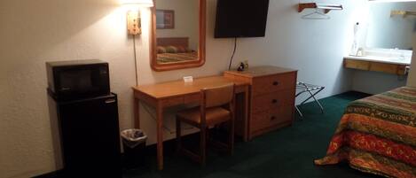 A hotel room with a desk, chair, and a bed. There is a television mounted on the wall, a mirror above the desk, and a small trash bin.