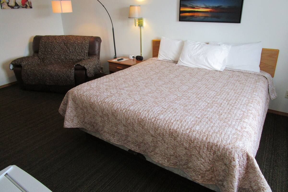 A hotel room with a large bed, a chair, a desk with a lamp, and a painting on the wall.