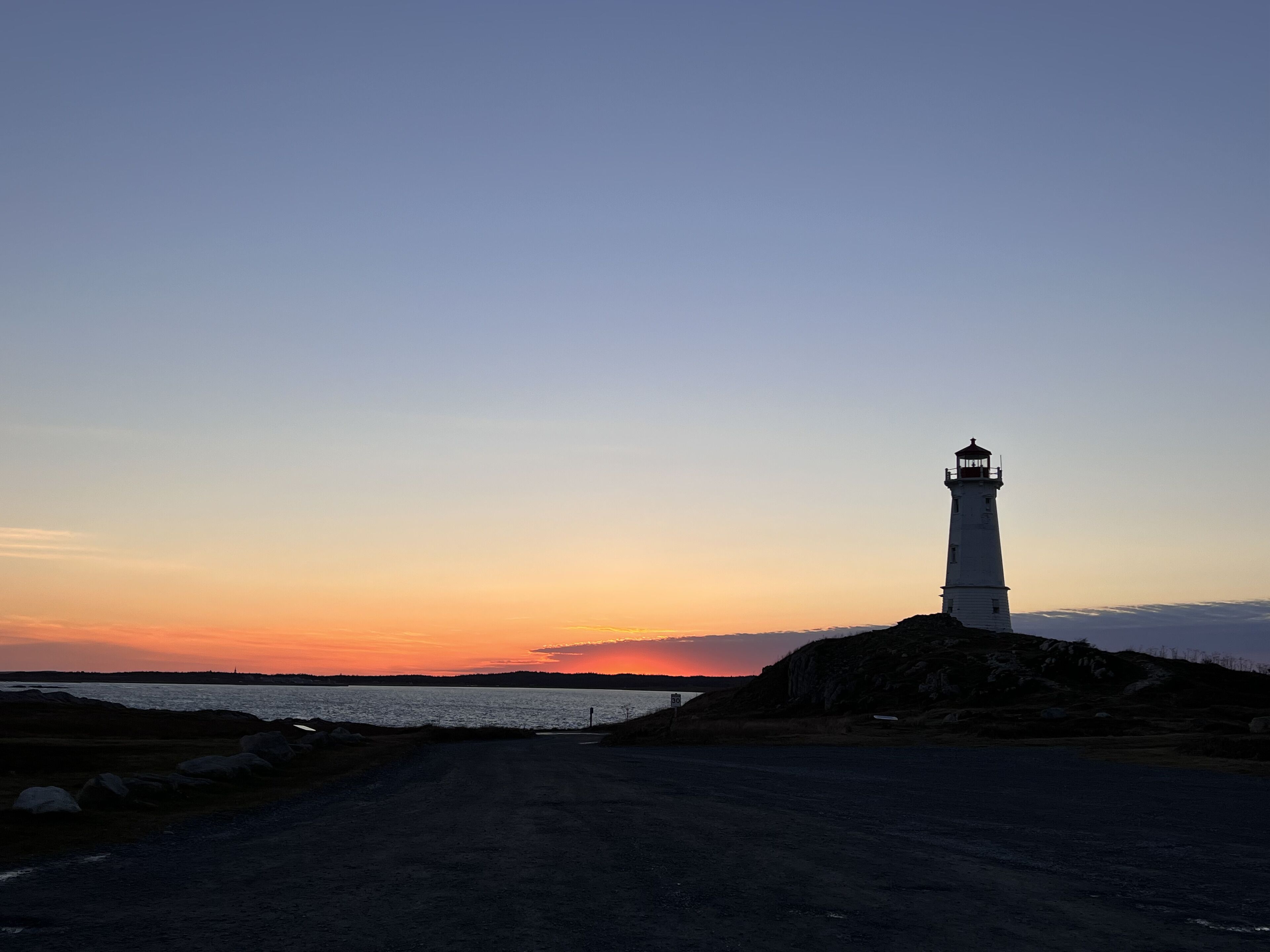 A lighthouse on a rocky outcrop with a clear sky and a sunset in the background.