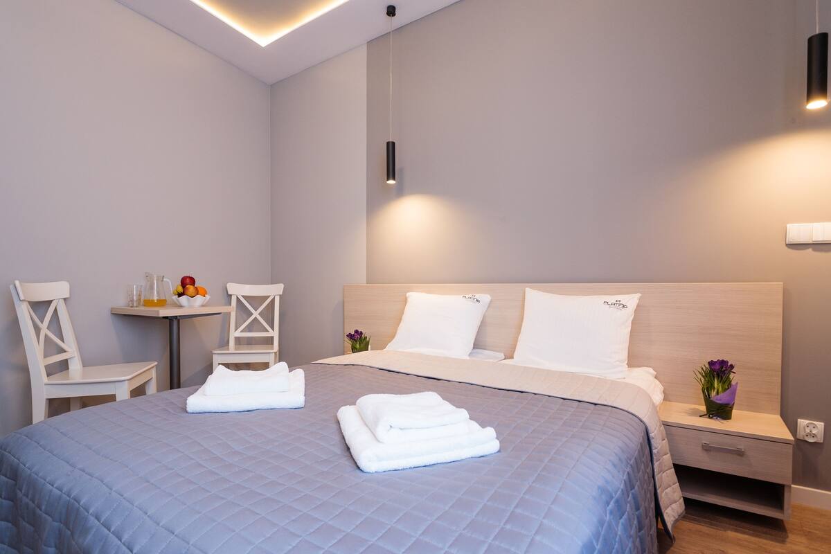 A modern hotel room with a large bed, a bedside table, a chair, and a small table with fruit and flowers.