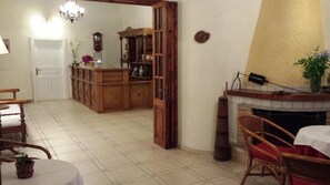 A traditional dining room with a wooden table, chairs, a fireplace, and a kitchen area with cabinets and a clock. - Ippokampos Hotel (Hydra)