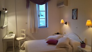 A bedroom with a bed, bedside tables, a chair, a window with curtains, and wall-mounted lamps. - Ippokampos Hotel (Hydra)