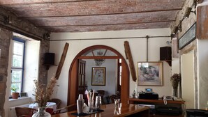 Ein rustikaler Raum mit freiliegenden Backsteinwänden, Holzbalkendecke und einem großen, gewölbten Durchgang. Es gibt einen Esstisch mit Stühlen, eine Vase mit Trockenblumen und ein gerahmtes Bild an der Wand.