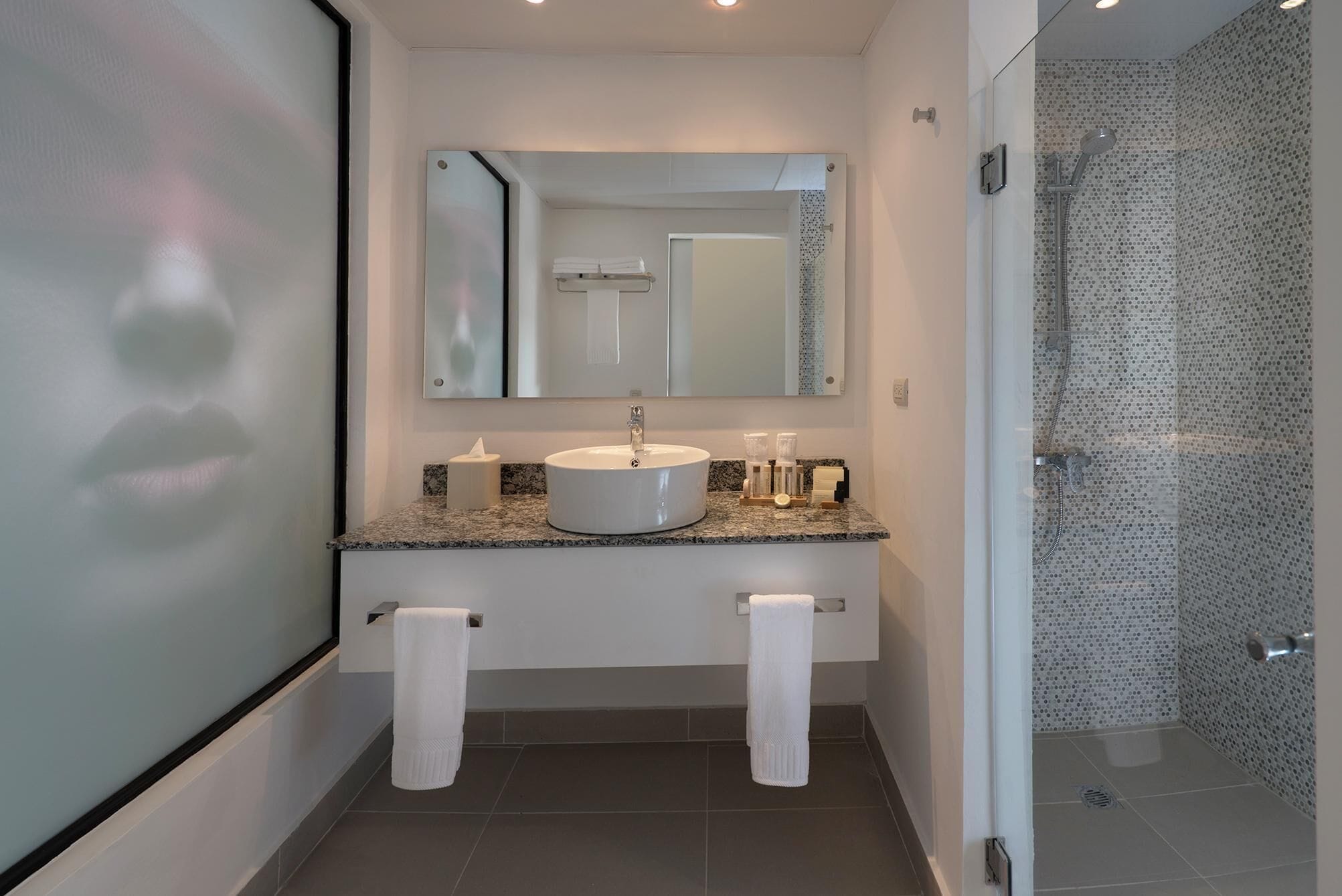 A modern bathroom with a granite countertop, a round sink, a large mirror, and a shower with a perforated glass door.