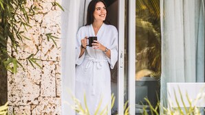 A person in a white robe standing at a glass door, holding a mug.