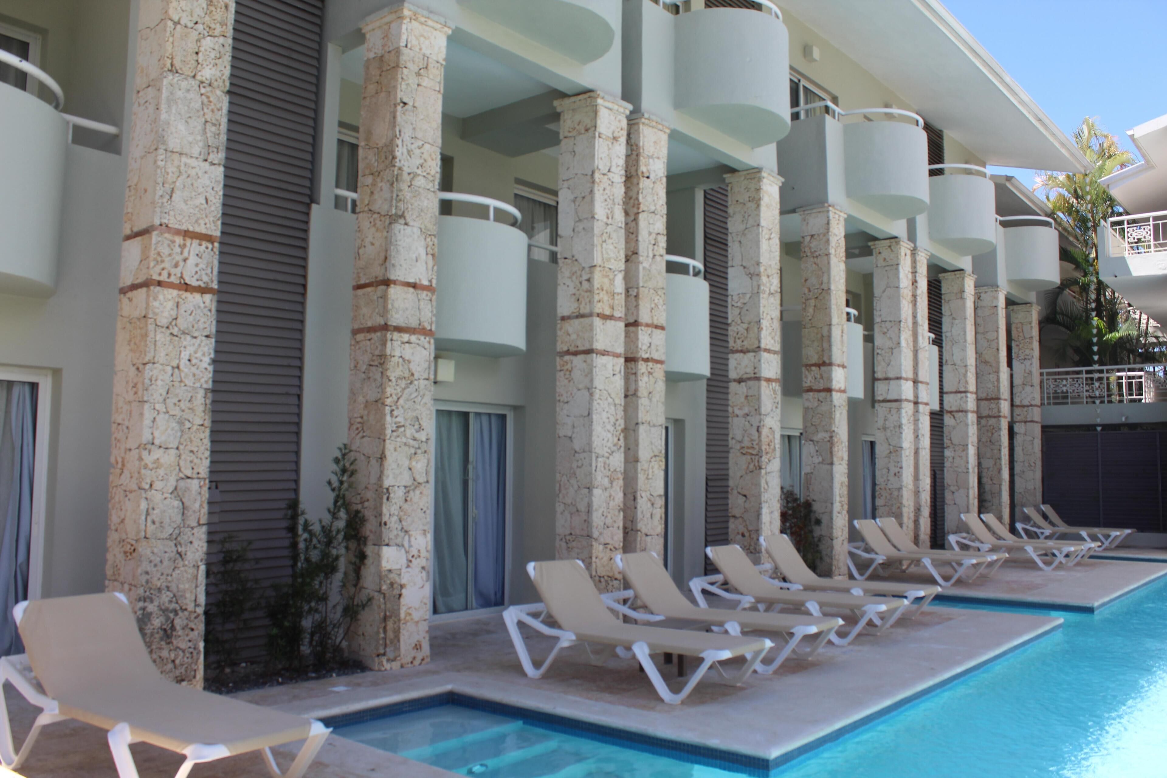 A poolside area with lounge chairs and a swimming pool, surrounded by stone pillars and a building with balconies.