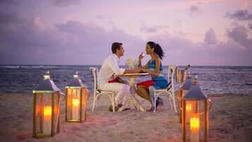 A couple dining at a table on a beach with lanterns and ocean in the background.