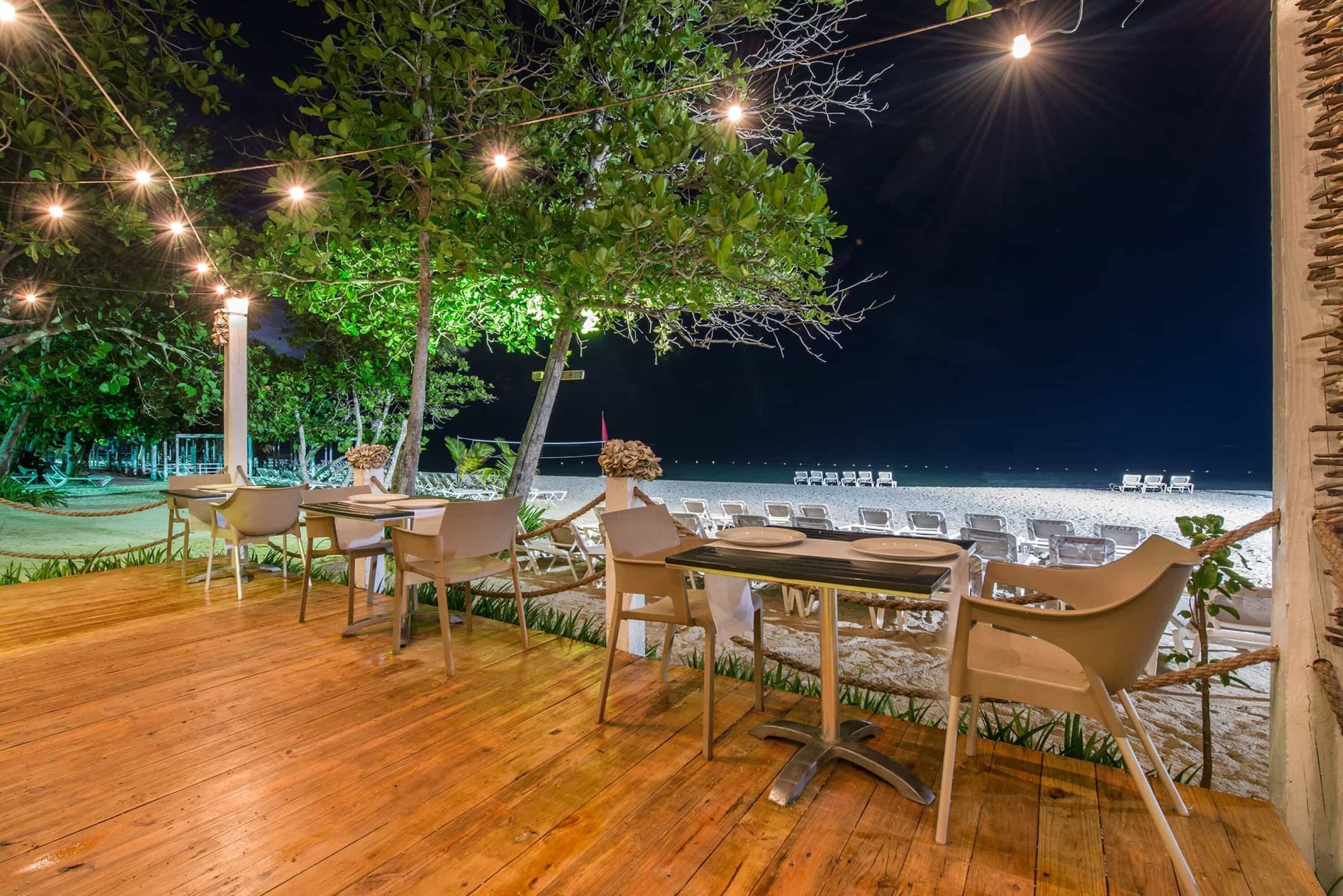 Un espacio de comedor al aire libre con piso de madera, mesas y sillas, con vista a una playa que tiene sillas de descanso y un bote.