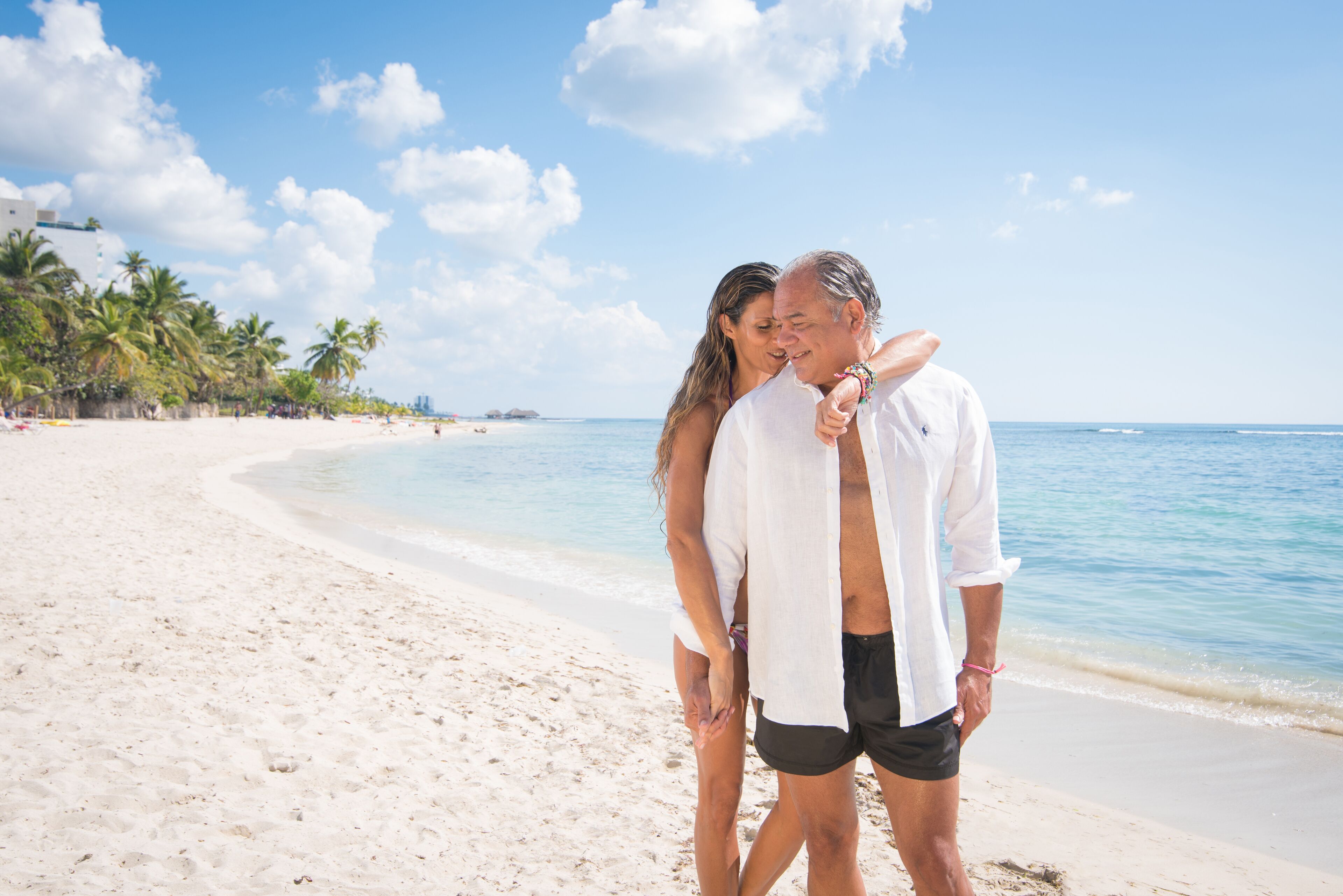 Une scène de plage où un couple se tient près l’un de l’autre, avec une plage de sable, des palmiers et un ciel bleu dégagé.
