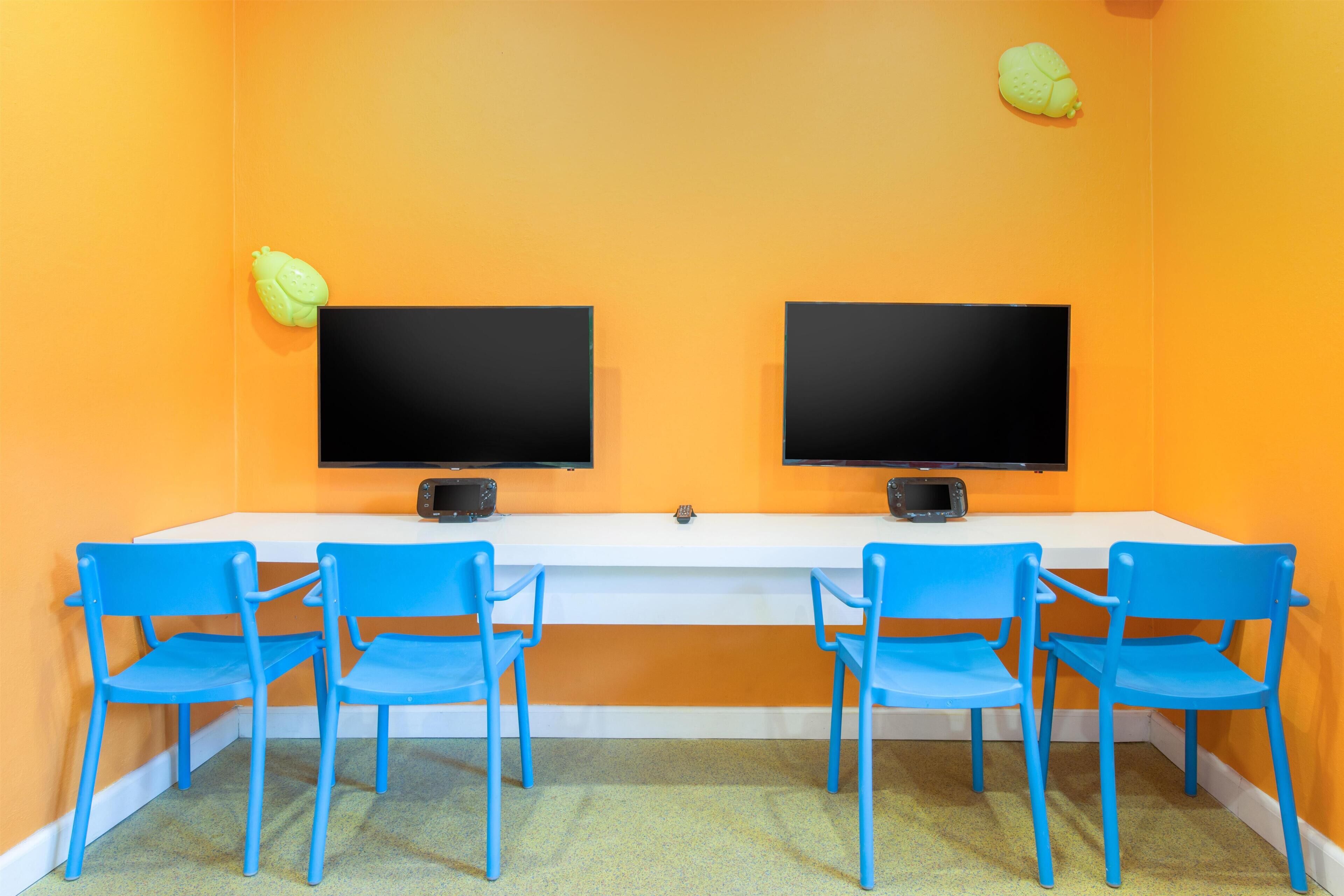 Um quarto com dois monitores pretos sobre uma escrivaninha branca, rodeado por quatro cadeiras azuis e paredes laranja.