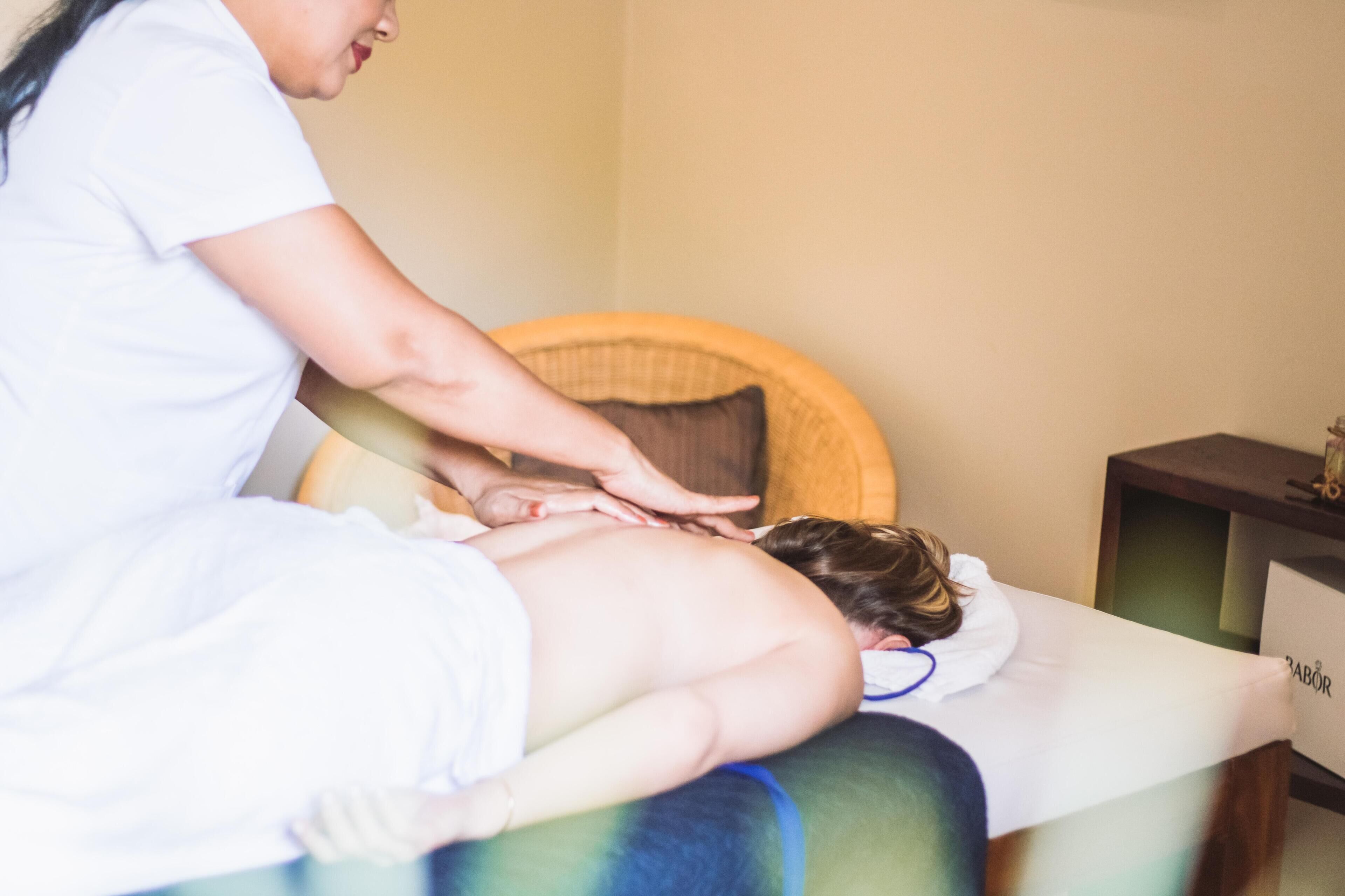 Une séance de massage dans une pièce équipée d’une table de massage, d’un fauteuil en osier et d’une petite table d’appoint en bois sur laquelle se trouve une bouteille.