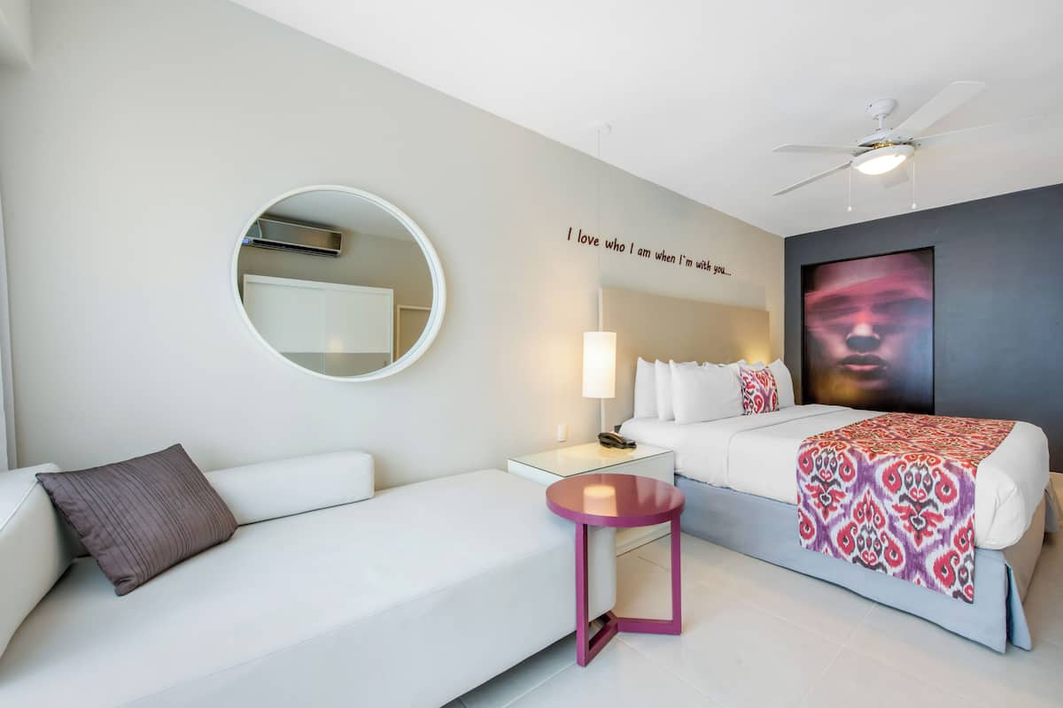 Ein modernes Hotelzimmer mit einem weißen Sofa, einem kleinen runden Tisch, einem Bett mit gemustertem Überwurf, einem runden Spiegel und einem Deckenventilator.