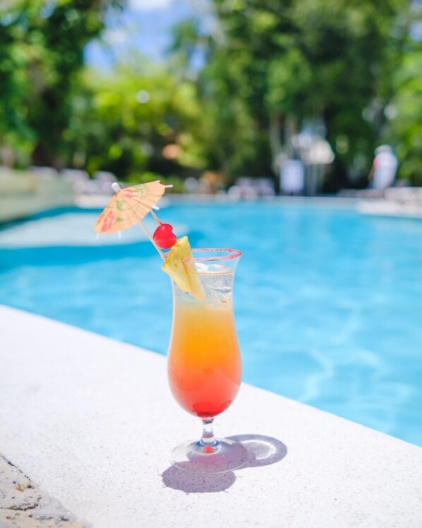 A cocktail with a cherry and a slice of pineapple, garnished with a small umbrella, placed on a poolside ledge.