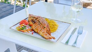 A dining table with a plate of food, a glass of white wine, and cutlery on a terrace with a view of the beach and ocean.