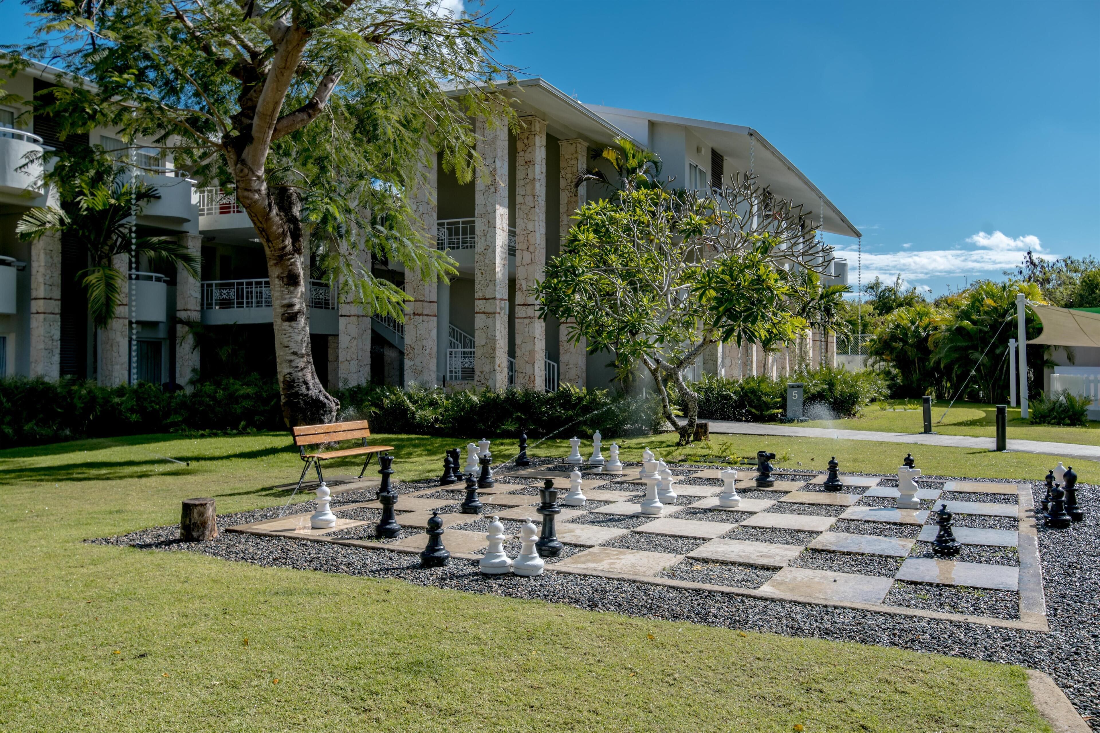 A chessboard set in a courtyard with a bench and trees, surrounded by a building with balconies and a clear sky.