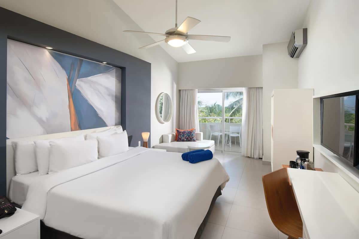 Ein modernes Hotelzimmer mit einem großen Bett, einem Deckenventilator, einem Fernseher und einem Balkon mit Blick auf Palmen.