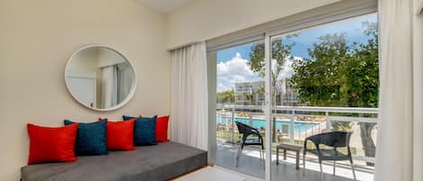 Une pièce moderne avec un canapé gris, des coussins rouges et bleus, un miroir rond et une vue sur une piscine et des bâtiments, visible à travers une porte vitrée.