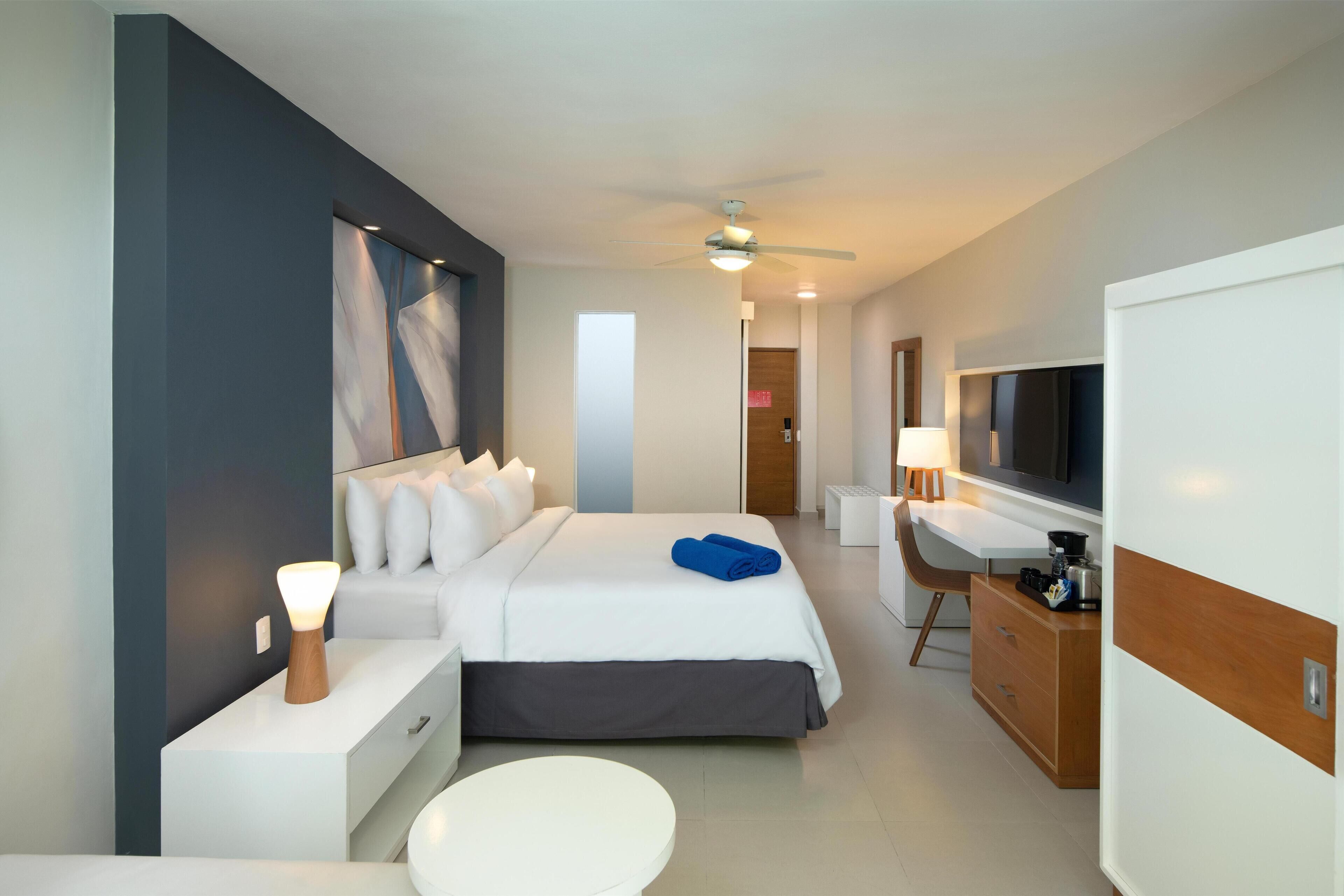 A modern hotel room with a bed, bedside table, lamp, TV, and a small desk.