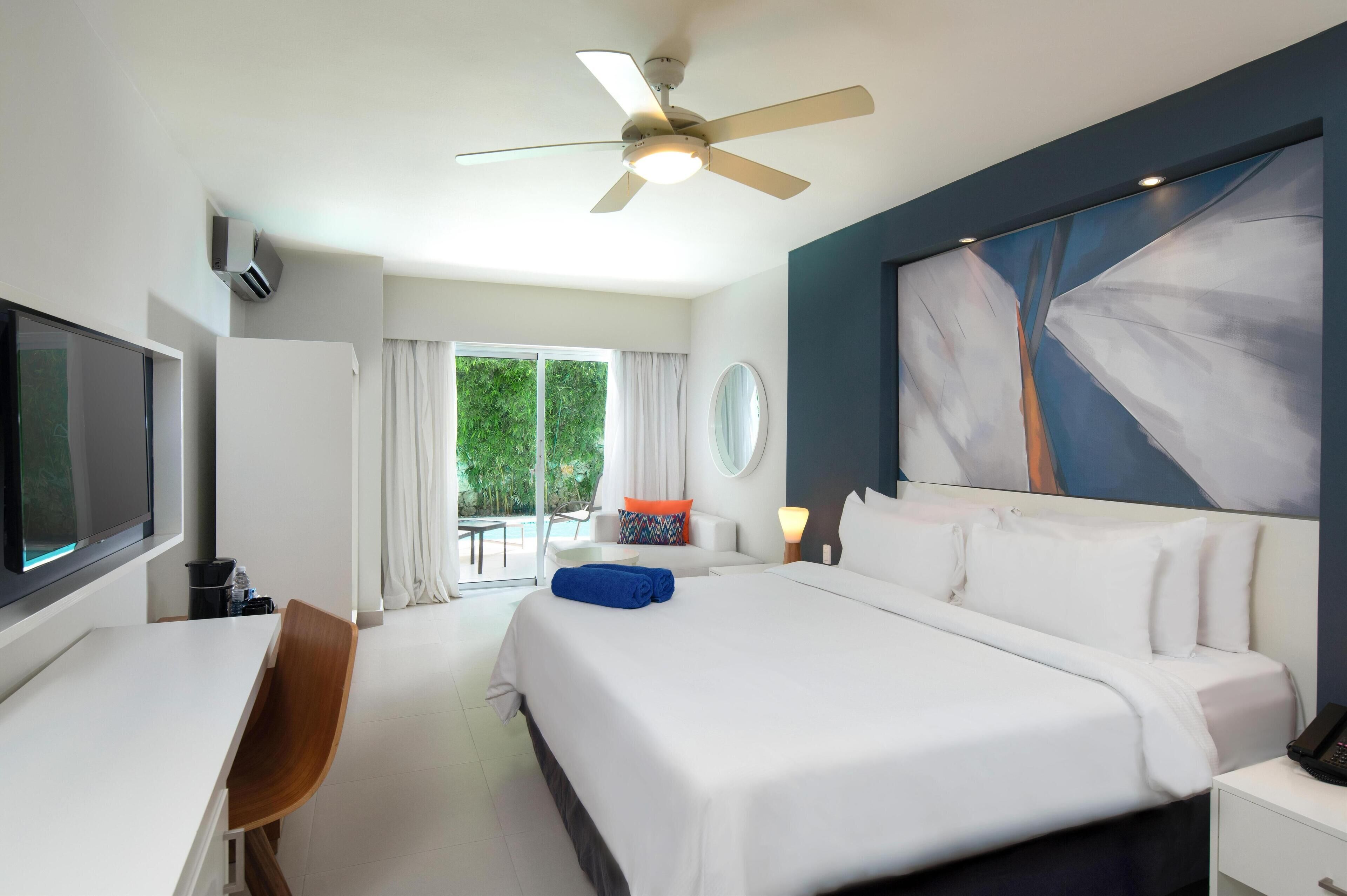 A modern hotel room with a large bed, a desk, a chair, a ceiling fan, and a large window with curtains.