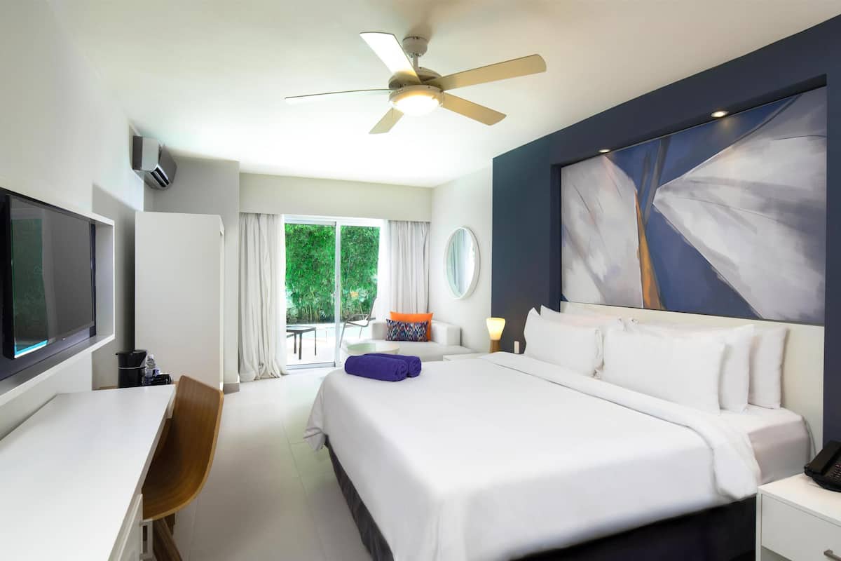 Ein Hotelzimmer mit Bett, Schreibtisch, Stuhl, Deckenventilator, Fernseher und einem Fenster mit Vorhängen.