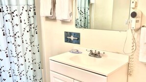 A bathroom with a white sink, a mirror, a towel rack, a shower curtain, and a trash bin. - Traveler's Inn Motel (Olney)