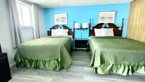 A hotel room with two beds, each with a green bedspread, a wooden headboard, and a nightstand with a lamp. There is a window with curtains, a wall-mounted painting, and a wooden panel on the right wall. - Traveler's Inn Motel (Olney)