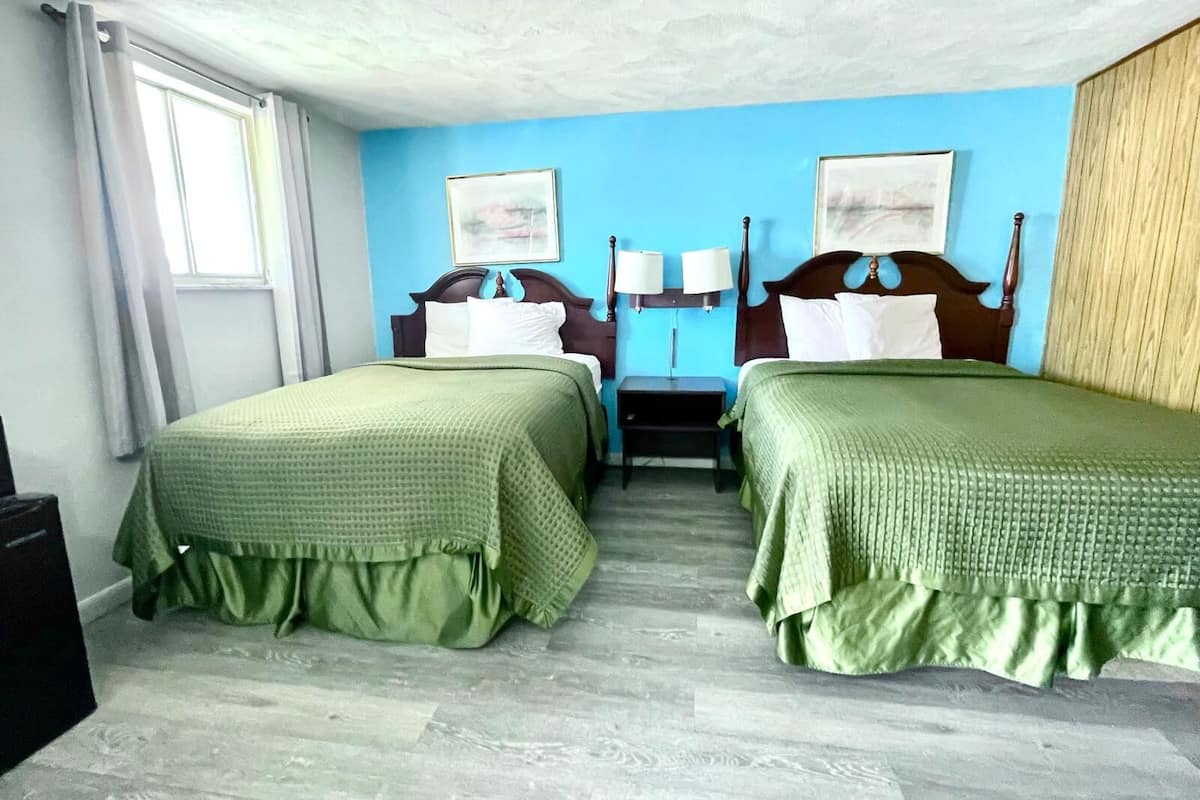 A hotel room with two beds, each with a green bedspread, a wooden headboard, and a nightstand with a lamp. There is a window with curtains, a wall-mounted painting, and a wooden panel on the right wall.