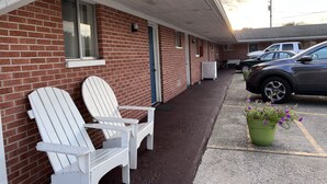 A motel with two white Adirondack chairs outside, a parking lot, and a green potted plant with purple flowers. - Traveler's Inn Motel (Olney)