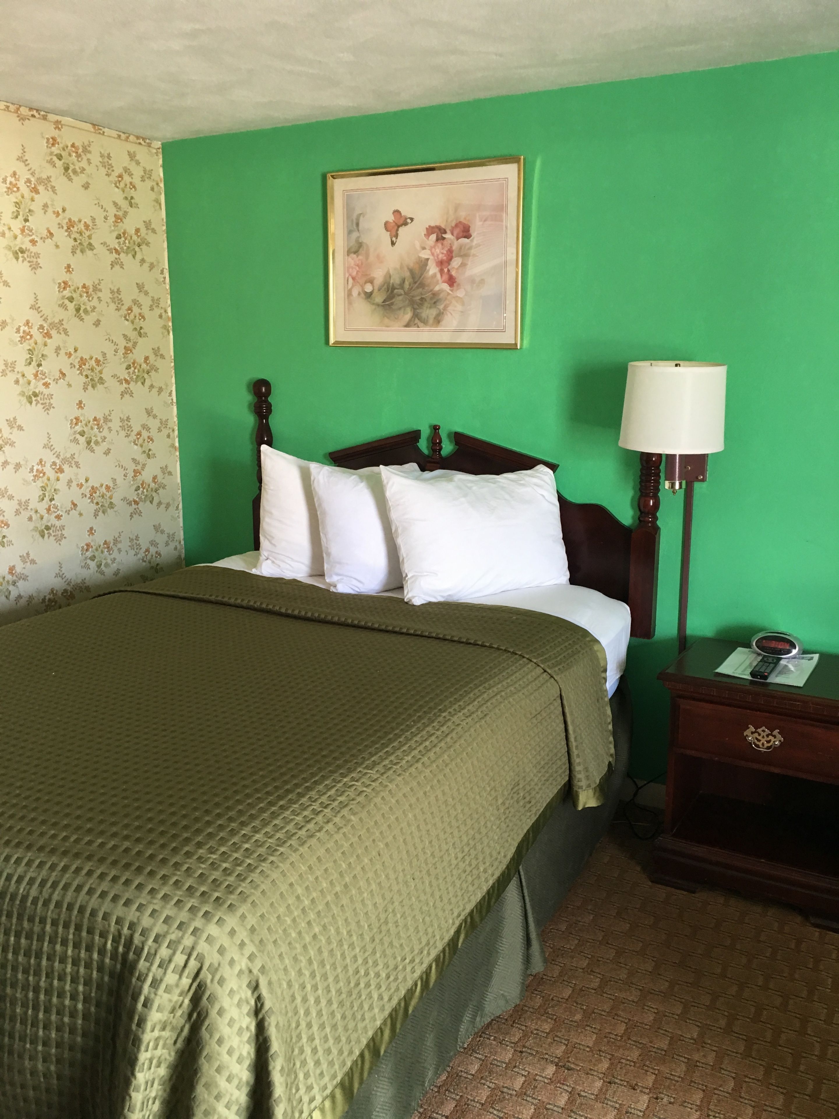 Una cama con colcha verde, almohadas blancas y cabecera de madera. En la pared, encima de la cama, hay un cuadro y una mesita de noche con una lámpara y un reloj.