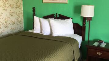 Una cama con colcha verde, almohadas blancas y cabecera de madera. En la pared, encima de la cama, hay un cuadro y una mesita de noche con una lámpara y un reloj.