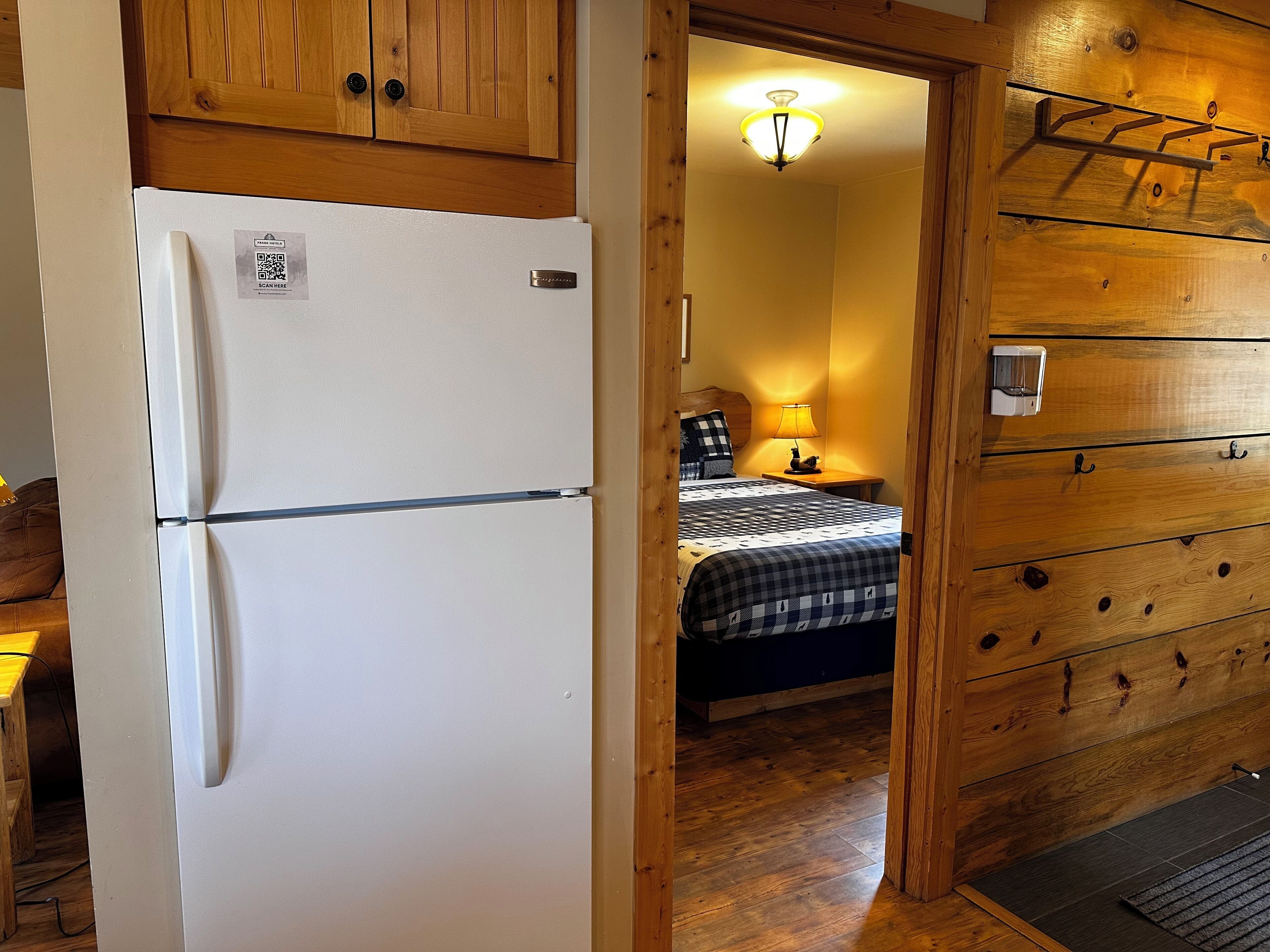 Une pièce de taille modeste, avec un réfrigérateur blanc, un lit recouvert d’une couverture à carreaux, une armoire en bois et une table de chevet avec une lampe.