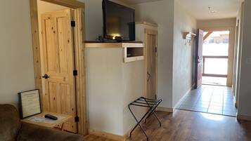 Una habitación con puertas de madera, un televisor sobre una estantería de madera, una mesita con un control remoto y un perchero metálico negro.