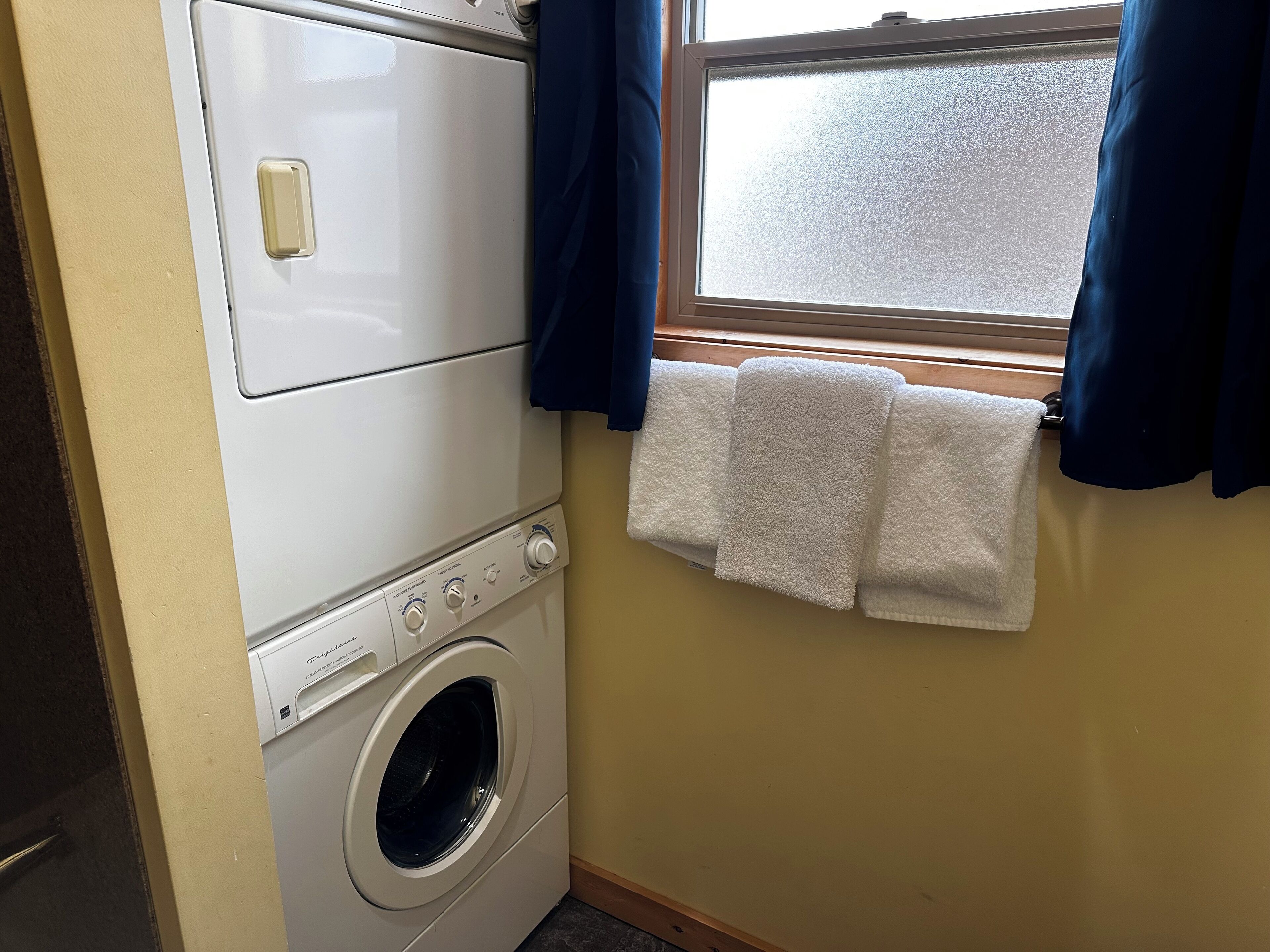 Un lavadero pequeño con lavadora y secadora de carga frontal, una ventana con cortinas y un perchero con dos toallas.