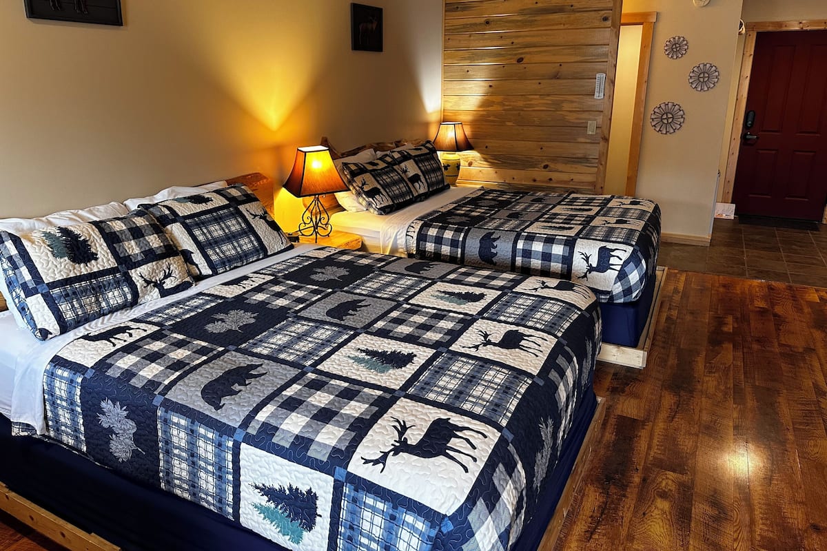A double bed with a checkered quilt featuring animal prints, two bedside lamps, a wooden headboard, and a door with decorative wall art.