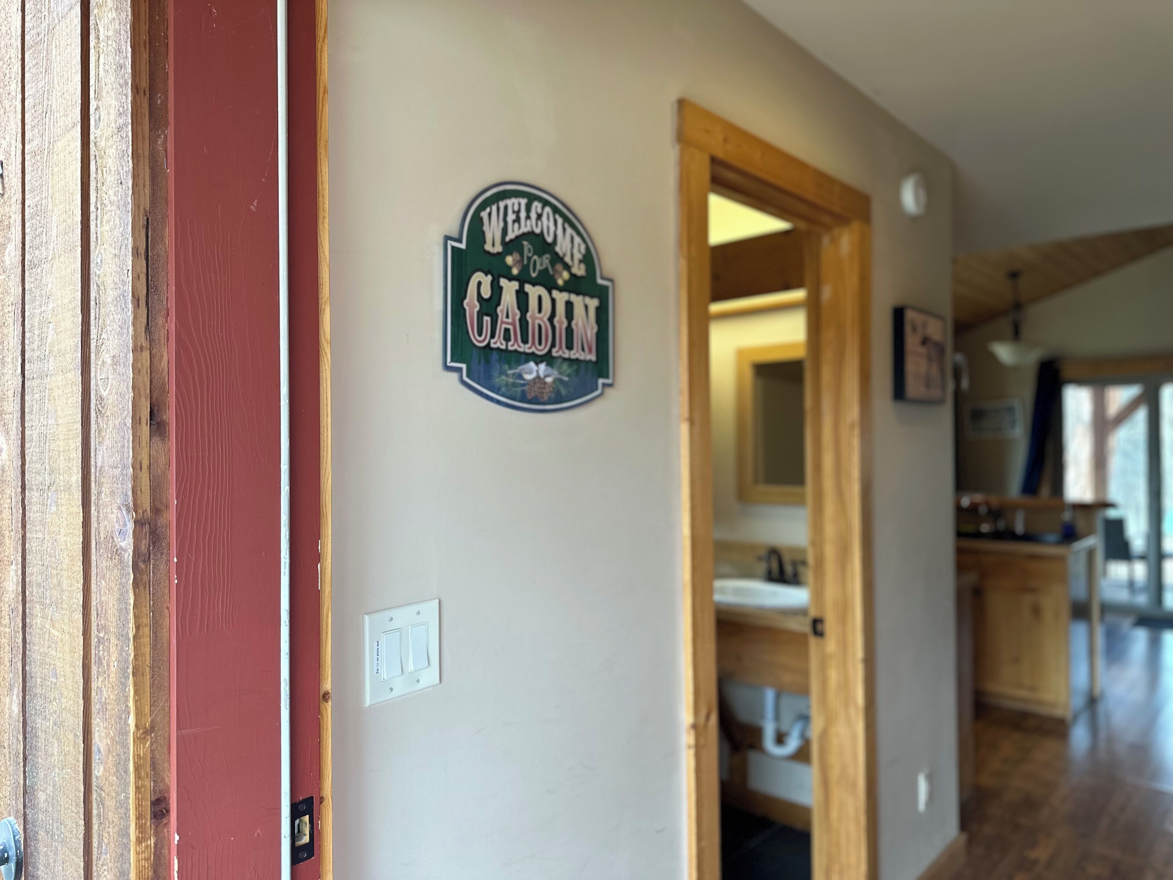 A hallway with a 'Welcome to Cabin' sign, a wooden door, and a glimpse of a bathroom with a sink and mirror.