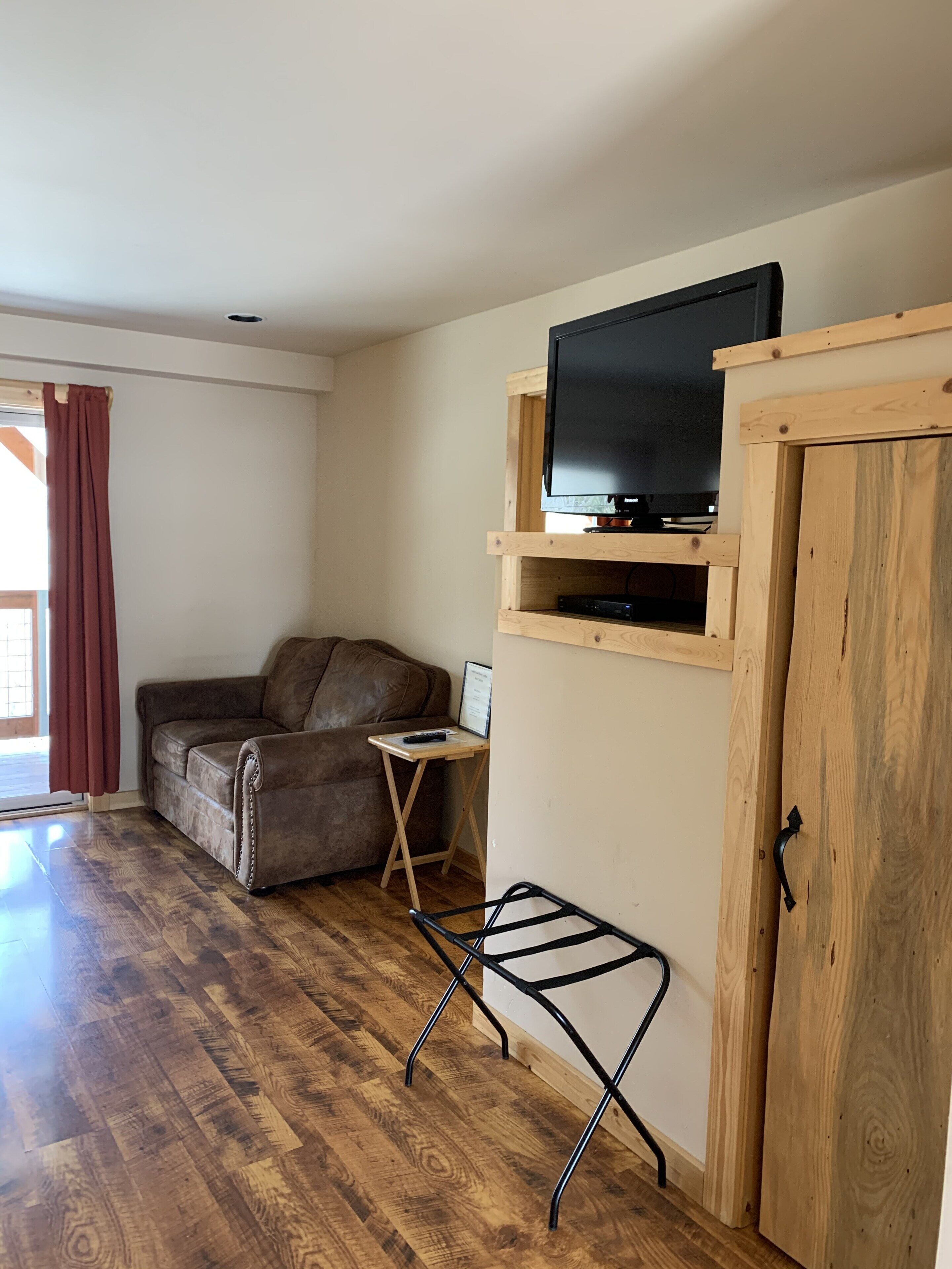 A living room with a wooden floor, a brown leather sofa, a television on a wooden shelf, and a small table with a remote control.