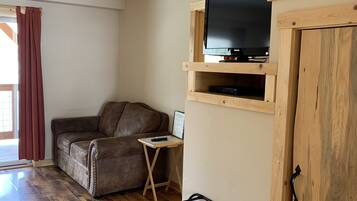 A living room with a wooden floor, a brown leather sofa, a television on a wooden shelf, and a small table with a remote control.