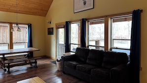 A cozy living room with a wooden ceiling, a black leather sofa, a wooden table, and a large window with curtains.